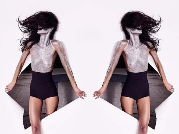 Glamorous mirrored shot of a slender woman covered in silver glitter, partially visible through torn white paper, set against a white backdrop.