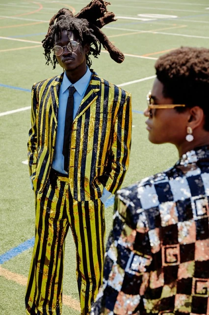 Two stylish young men in patterned suits pose on a green field. One wears a gold and black striped suit, and the other a black and white checked suit.