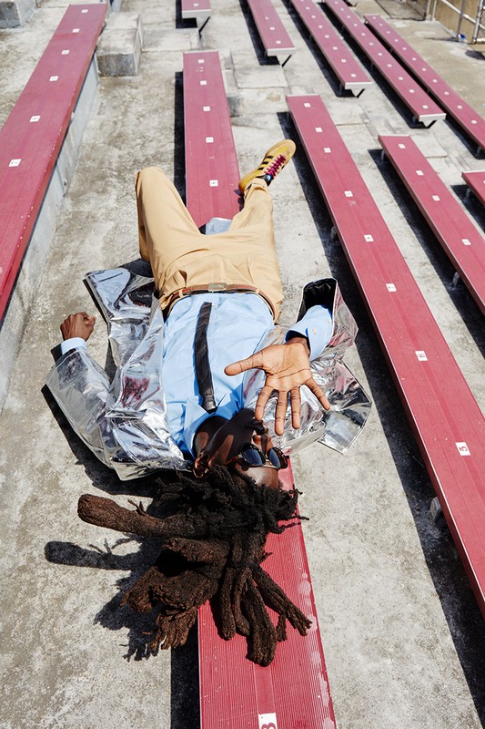 A person with dreadlocks and sunglasses lies on a red stadium bench, wearing a light blue shirt, tan pants, and a silver jacket.