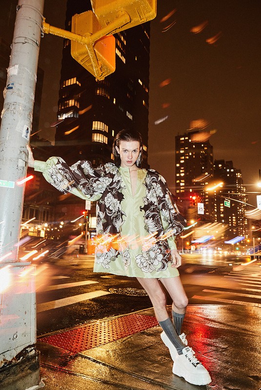 Fashion model in an ethereal green floral dress poses on a city street at night, leaning against a pole with traffic lights.