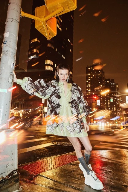 Fashion model in an ethereal green floral dress poses on a city street at night, leaning against a pole with traffic lights.