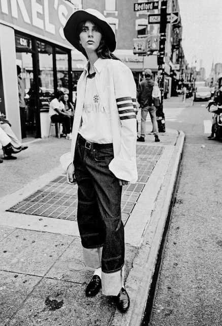 A woman in a hat, jeans, and oversized shirt standing on a city sidewalk, with buildings and a store in the background in black and white.