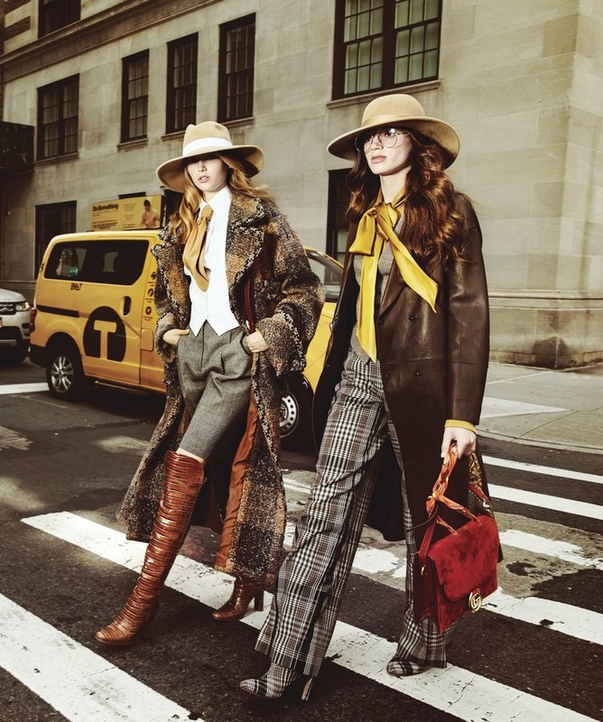 Two women in fashionable coats and hats walk across a crosswalk in an urban setting, next to a yellow taxi cab and buildings.