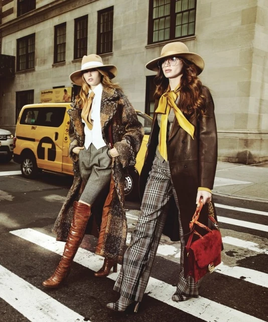 Two women in fashionable coats and hats walk across a crosswalk in an urban setting, next to a yellow taxi cab and buildings.