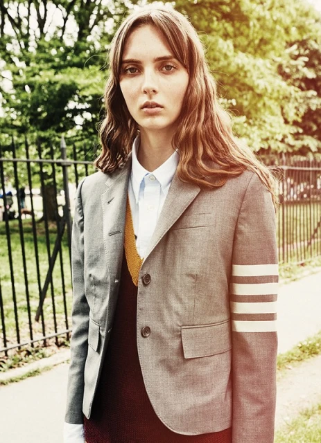 Stylish young woman in grey blazer, shirt, and vest poses outdoors with a neutral expression; trees and wrought iron fence in background.