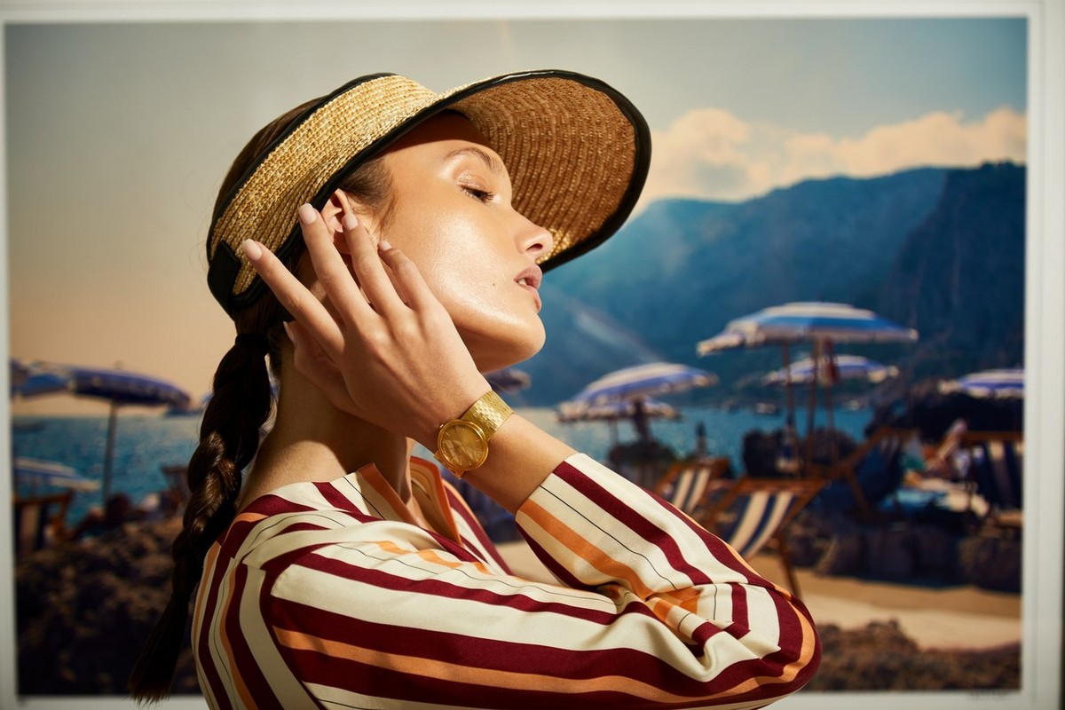 Woman in straw visor poses with hand near face, wearing striped top and gold watch, set against beach landscape backdrop indoors.