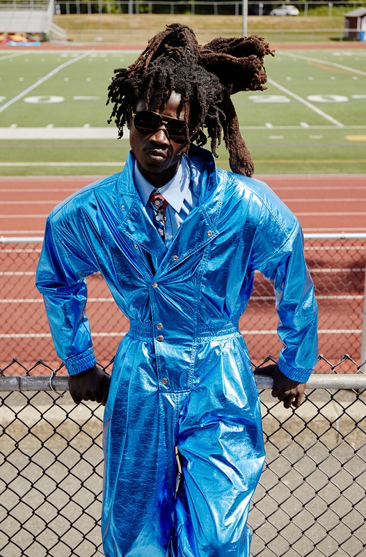 A man with dreadlocks and sunglasses is leaning against a fence in a shiny blue jumpsuit, posing in front of a green sports field.