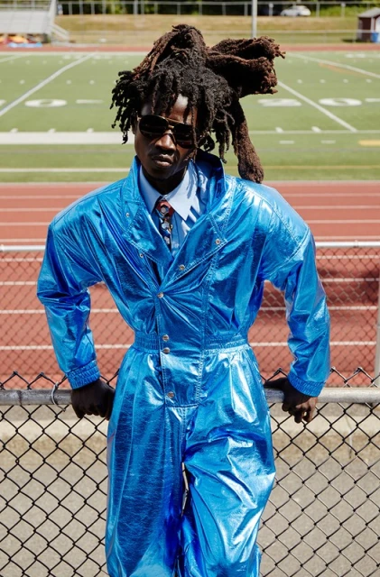 A man with dreadlocks and sunglasses is leaning against a fence in a shiny blue jumpsuit, posing in front of a green sports field.