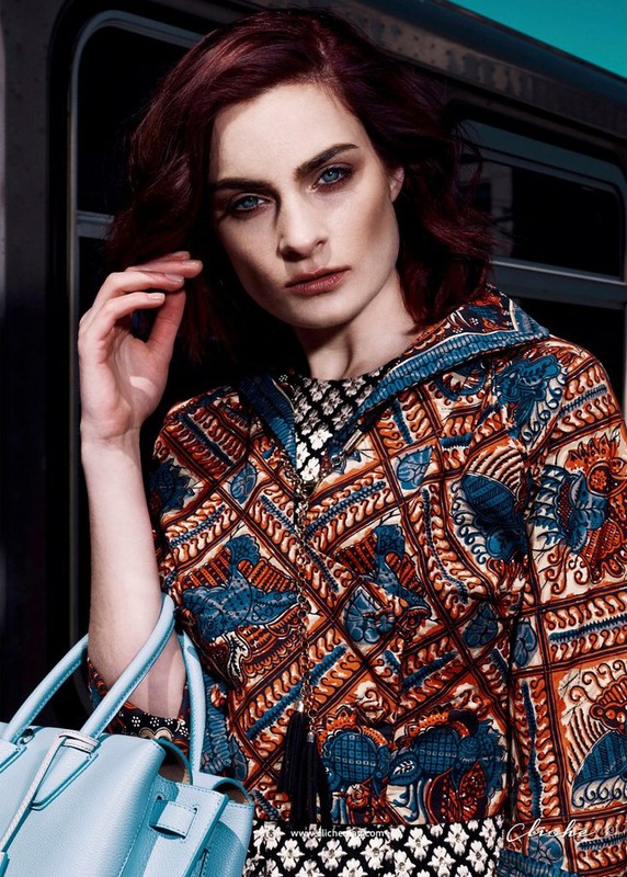 A red-haired woman in a patterned jacket holds a light blue purse in an indoor setting with teal and black backgrounds.