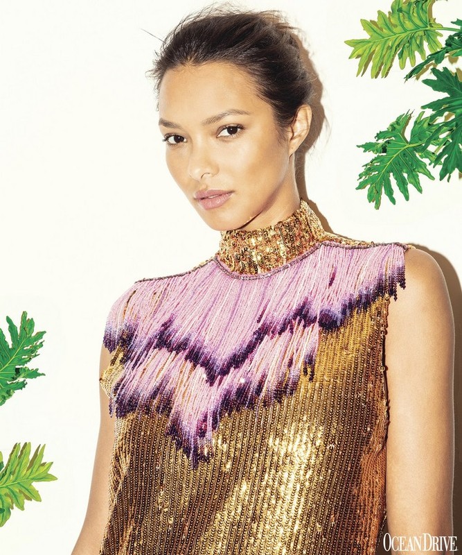 Portrait of a woman with fair skin and dark hair wearing a gold sequined dress with pink fringe against a neutral backdrop.