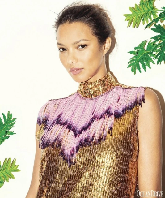 Portrait of a woman with fair skin and dark hair wearing a gold sequined dress with pink fringe against a neutral backdrop.