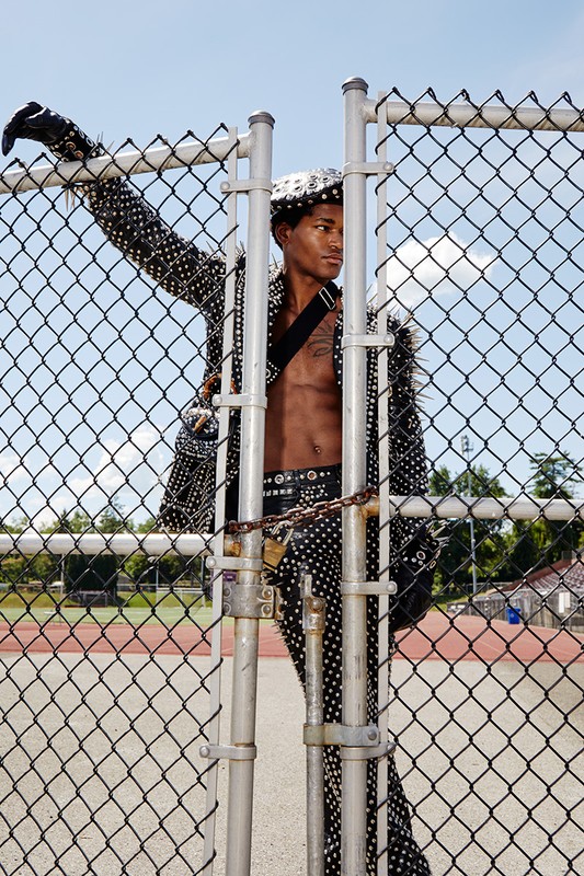 A young, shirtless African American man poses against a chain-link fence in a studded jacket, belt, and hat. The setting is an outdoor track.