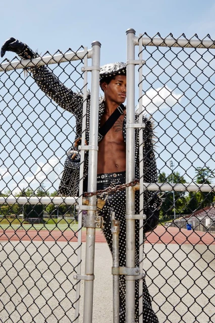 A young, shirtless African American man poses against a chain-link fence in a studded jacket, belt, and hat. The setting is an outdoor track.