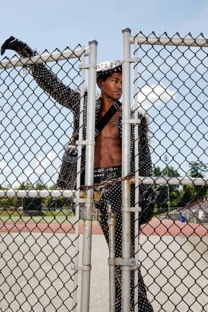 A young, shirtless African American man poses against a chain-link fence in a studded jacket, belt, and hat. The setting is an outdoor track.
