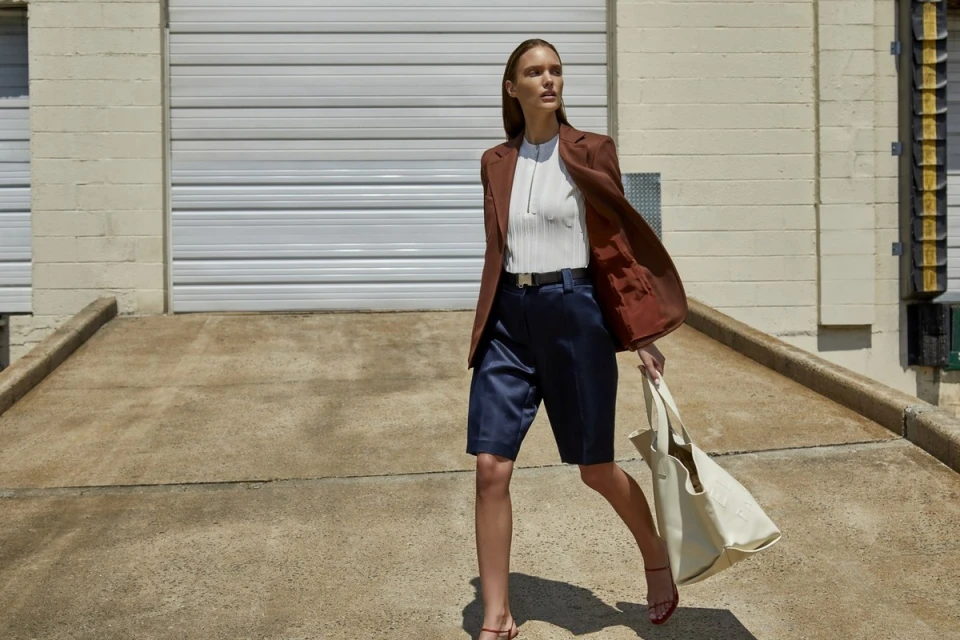 Adult woman strides confidently outdoors in a rust-colored blazer and navy shorts, holding a beige tote bag against a neutral backdrop.