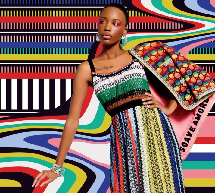 A Black female fashion model poses in a multicolor knit dress against an abstract colorful background, accessorized with earrings and bracelets.