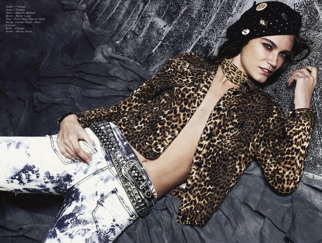 Fashion shot of a woman in an unbuttoned leopard-print jacket, beret, and acid-wash jeans, reclining against a distressed gray backdrop.