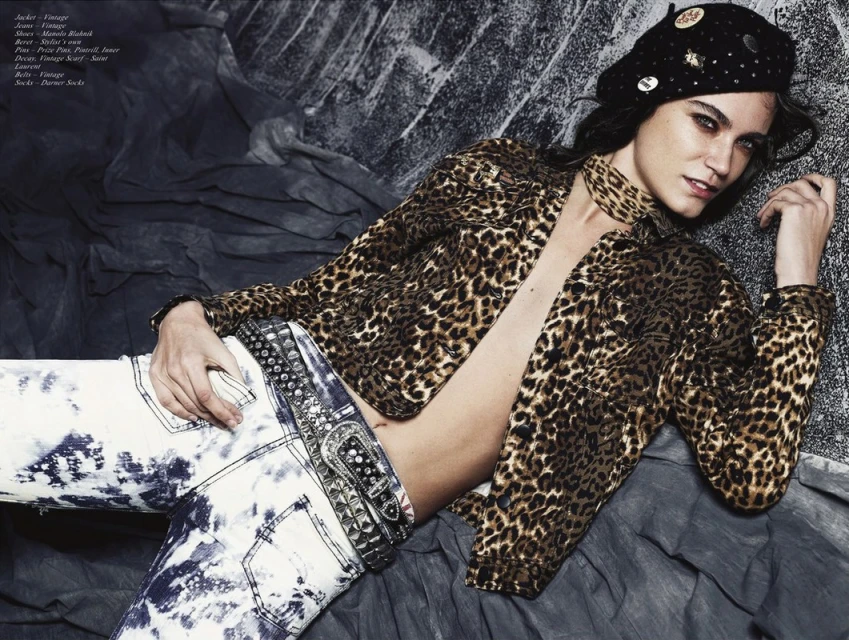 Fashion shot of a woman in an unbuttoned leopard-print jacket, beret, and acid-wash jeans, reclining against a distressed gray backdrop.