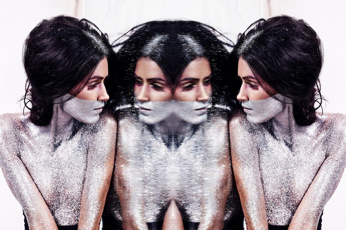 Woman with dramatic silver glitter makeup in a studio photo, mirrored reflections on either side, creating a surreal, beauty-focused aesthetic.