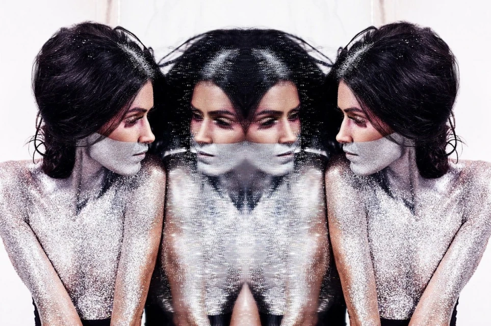 Woman with dramatic silver glitter makeup in a studio photo, mirrored reflections on either side, creating a surreal, beauty-focused aesthetic.
