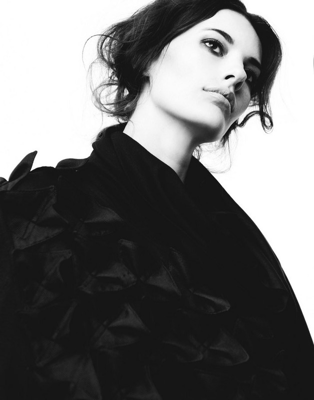 Monochrome portrait of a stylish woman with dark hair, wearing a black ruffled coat, against a bright white background in a studio setting.
