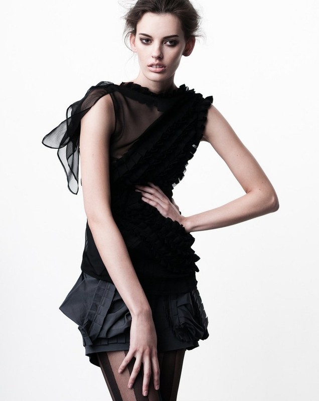 Woman in black ruffled top and grey shorts poses in a fashion studio portrait with a bright white backdrop and soft, diffused lighting.