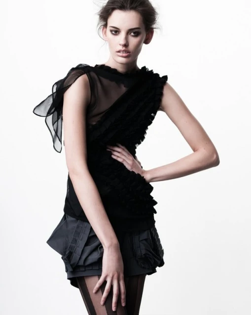 Woman in black ruffled top and grey shorts poses in a fashion studio portrait with a bright white backdrop and soft, diffused lighting.