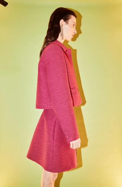 A woman in profile poses in a magenta jacket and skirt, accessorized with dangling earrings, against a bright chartreuse background.