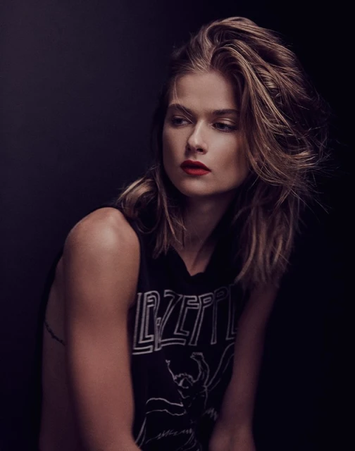 Portrait of a young adult female with blonde hair and red lipstick wearing a Led Zeppelin sleeveless top in front of a dark backdrop.