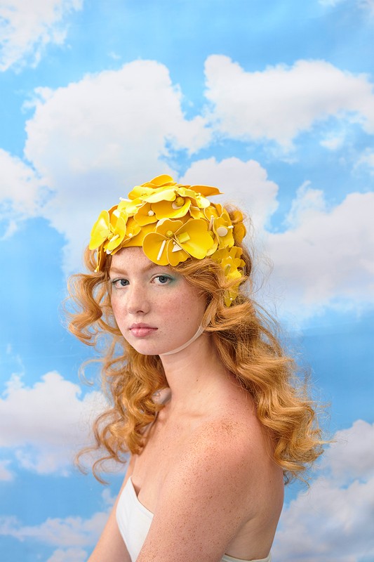 Red-haired woman wearing a yellow flower headdress and white dress, posing in front of a background featuring a blue sky and white clouds.