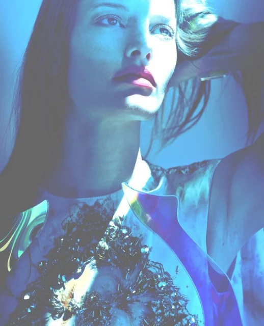 Close-up of a fashion model with dramatic blue lighting, accented by vibrant pink lipstick, wearing a decorative, iridescent top.