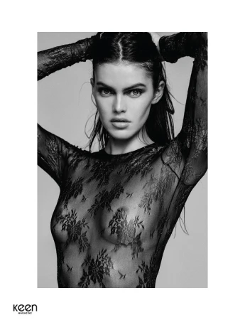 Black and white portrait of a woman wearing a sheer black lace top with her arms raised behind her head.