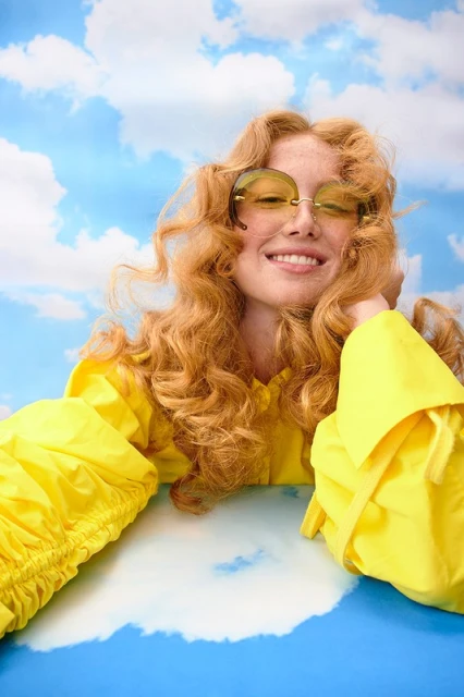Woman with red hair and sunglasses smiles in front of a backdrop with a blue sky and clouds while wearing a yellow jacket.