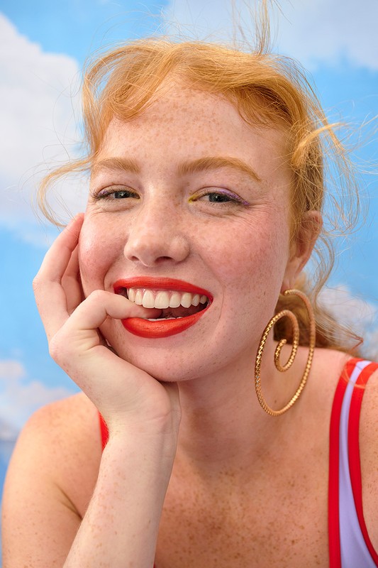 Close-up portrait of smiling redhead woman with freckles, bright red lipstick, large gold earrings, and a blue sky and cloud background.