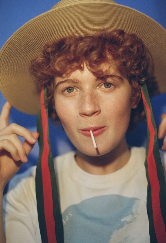 Young adult with curly red hair wears a straw hat with ribbons, a graphic tee, and a lollipop against a light blue background.