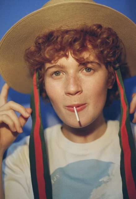 Young adult with curly red hair wears a straw hat with ribbons, a graphic tee, and a lollipop against a light blue background.