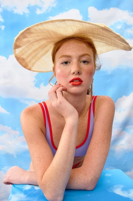 Portrait of a freckled woman with an orange lipstick wearing a sun hat and a purple swimsuit against a sky background in a studio.