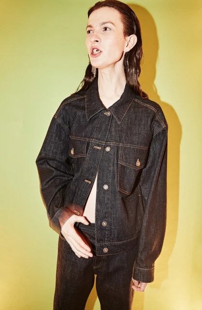 A young adult female poses in a dark denim jacket and jeans against a gradient yellow-green background in a studio fashion shot.