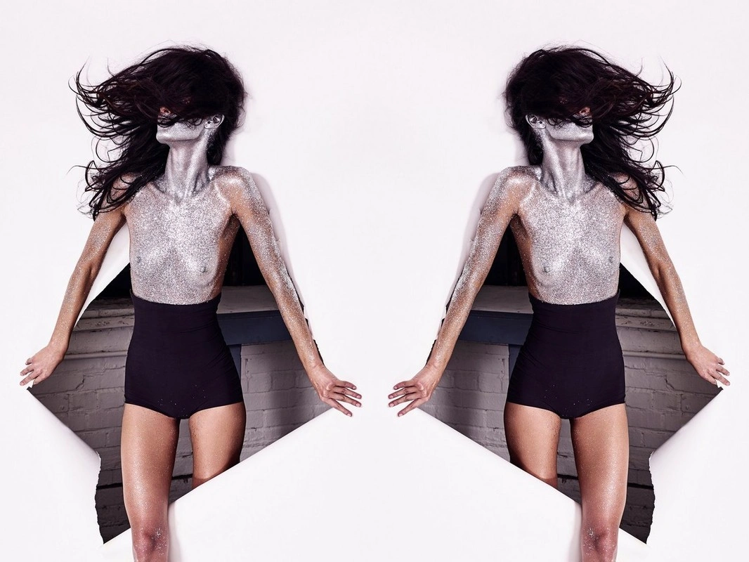 Model with silver glitter body paint and dark hair blowing back, emerging through a torn white paper backdrop with black shorts