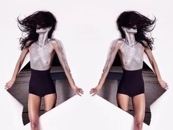 Model with silver glitter body paint and dark hair blowing back, emerging through a torn white paper backdrop with black shorts