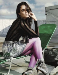 Woman in sheer black striped top and metallic skirt sitting on a green chair under a cloudy sky.