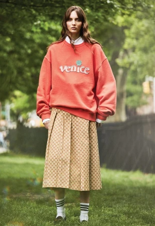 Model in red sweatshirt and monogram skirt standing on a park lawn with trees in Los Angeles