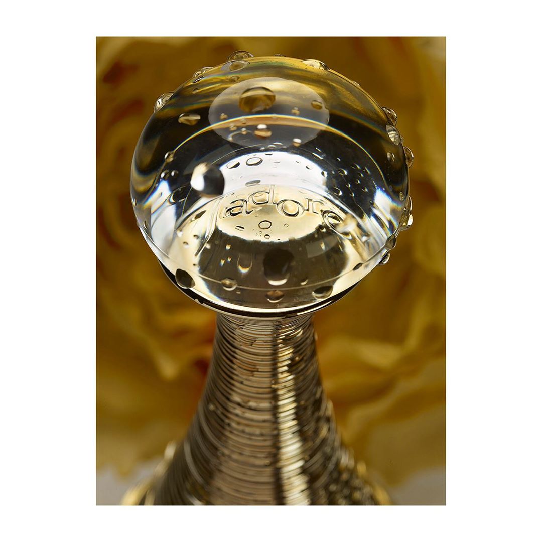 Close-up studio shot of a clear glass perfume bottle with water droplets and a gold metal neck, set against a soft yellow floral background.