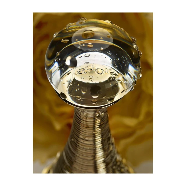 Close-up studio shot of a clear glass perfume bottle with water droplets and a gold metal neck, set against a soft yellow floral background.