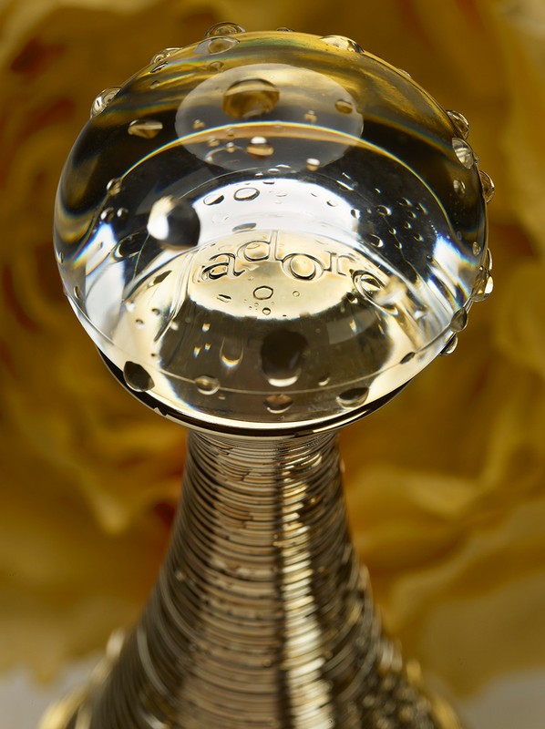 Close-up photo of a gold perfume bottle with a clear glass sphere on top, featuring the text 'J'ADORE,' set against a blurred yellow backdrop.