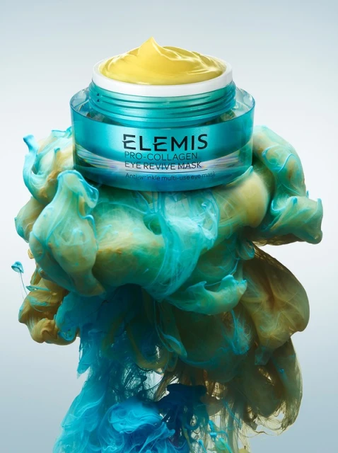 Elemis Pro-Collagen Eye Revive Mask sits atop swirling blue and yellow pigment clouds against a gradient light backdrop.