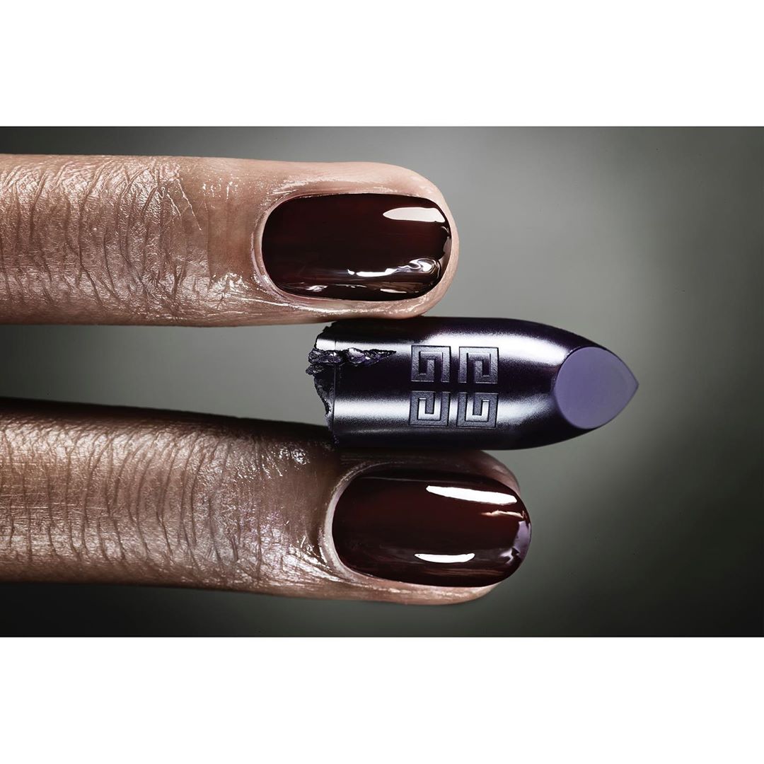 Close-up product shot of Givenchy lipstick bullet held between two manicured fingers with dark, glossy nail polish and a gray gradient background.