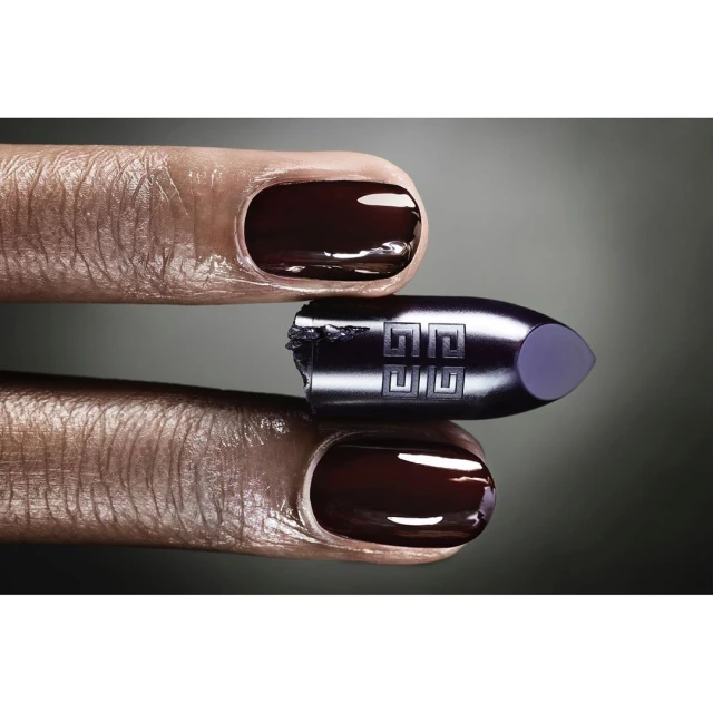 Close-up product shot of Givenchy lipstick bullet held between two manicured fingers with dark, glossy nail polish and a gray gradient background.