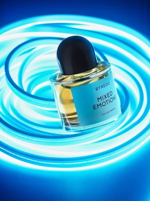 Byredo Mixed Emotions Eau de Parfum bottle on blue gradient background with neon-blue light accents. Product shot for beauty advertisement.