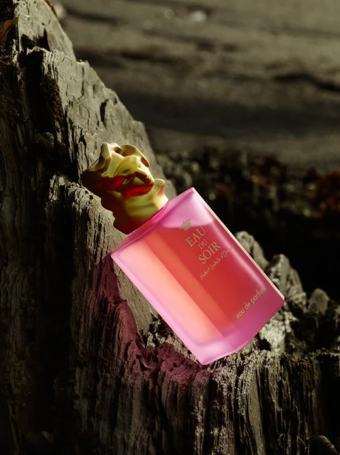 Elegant studio shot of a perfume bottle with a gold cap on a rock, showcasing its pink glass and contrasting textures in dim lighting.